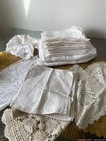 Vintage white table linens including folded napkins, tablecloths, and a round dinner roll holder with lace trim, displayed on a table.