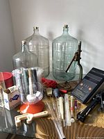 Glass carboys, wine making accessories, PH tester and bottle capper setup shown together on a table.