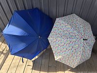Two large golf umbrellas standing opened on a wooden floor, one navy blue and the other white with colorful heart designs.