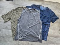 Photo showing front side of three Under Armour men's shirts in size XL: olive green short sleeve, grey sleeveless, navy blue short sleeve with camo sides.