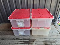 Four clear plastic storage bins stacked in two pairs, each with red lidded tops, placed on wooden floor against metal wall.