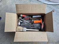 Open cardboard box packed with various masonry tools including trowels, chisels, brushes and other items showing wear and use