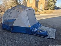 Tent fully assembled showing front side with extended entry flap on gravel surface. Grey and blue fabric with black and orange poles visible.