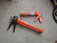 Two orange caulking guns, one smaller and one larger, placed on a concrete floor