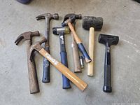 Photo showing seven assorted hammers and mallets laid on concrete floor, displaying handles and heads clearly.