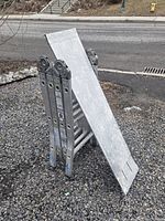 Folded multi-position aluminum ladder standing on gravel surface with work platform attached top side.