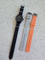 Top view of watch on black/grey ZuluDiver NATO strap next to two extra silicone straps in grey and orange laid out on countertop.