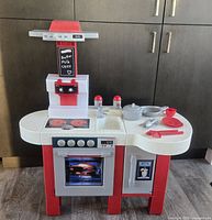 Full view of the Miele Kids Play Kitchen set with accessories arranged on countertop: stovetop with two burners, oven door closed, microwave, sink, salt and pepper shakers, cooking utensils, plate, cup, fork, knife, spoon.