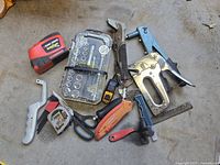 Full view of lot of hand tools on concrete floor including staple guns, riveter, tape measures, ratchet set, scissors, utility knife, hammer, file.