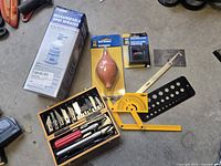 Photo of the lot showing the X-Acto cutter set, rechargeable spot sprayer box, battery tester in packaging, dust blower/soufflette in packaging, and additional measurement and drafting tools.