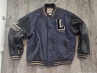 Front view of vintage Levi's varsity jacket showing navy wool body, black leather sleeves, and embroidered patches including large 'L' on chest and 'Levi Strauss' on sleeve, with striped ribbed collar, cuffs, and hem.