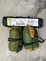 Coleman 4-person instant cabin tent in black carry bag and two green/yellow sleeping bags in compression sacks on concrete floor.