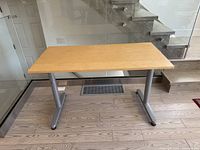 Front view of IKEA adjustable height desk with wooden top and silver metal legs on wood patterned floor near glass panel and stairs.