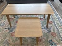 Matching coffee table and side table set with light ash veneer tops and solid birch legs placed on floral patterned rug in front of gray couch.