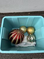 Blue bin containing four large Christmas ornaments: one large red and gold striped ornament, one gold glitter ornament, one gold matte ornament, and one shiny green ridged ornament.