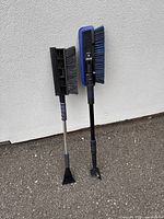 Two extendable car snow brushes side by side resting against a wall on asphalt surface, showing full length and brush and scraper ends.