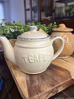 Front and side views of the cream-colored ceramic teapot with embossed 'TEAPOT' and blue floral pattern on lid knob, next to a brown-orange ceramic teapot.