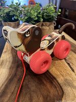 Photo showing front and side view of the Fisher Price Snoopy dog pull toy with red wheels on wooden surface and background plants.