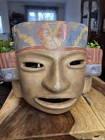 Front view of handcrafted pottery mask with colorful geometric and tribal designs on headdress, carved clay face details, and side ear ornaments.