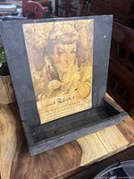 Metal bottle cap catcher with a vintage Coca Cola advertisement of a woman holding a bottle, showing signs of wear and aging.