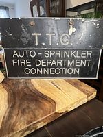 Front view of the vintage TTC metal fire department connection sign resting on a wooden surface with visible wear and rust spots.