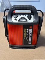 Front view of the Black & Decker jump starter showing orange sides, black handle, and front control panel with power switch and LED indicators.