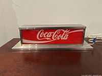 Front view of the rectangular Coca-Cola sign showing the red background with white script logo and metal frame base
