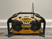 Frontal view of yellow and black DeWalt job site radio with protective black tubular frame, dual speakers, radio display, and controls visible. Surface shows usage wear.