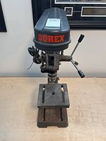 Front view of Durex bench top drill press showing the top cover with red logo, drill chuck, handle, and work surface with slotted grooves, mounted on a heavy base.