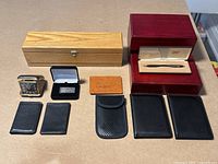 Lot displayed on cardboard with all items visible including leather wallets, travel clock, money clip, pen, and wooden boxes.