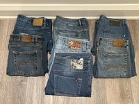View of all seven jeans pairs folded and laid out showing different denim washes and brand patches on the waistbands