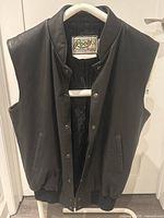 Front view of black leather vest on hanger showing snap-button closure and ribbed waistband.