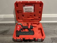Milwaukee 18-gauge pneumatic brad nailer placed inside open red molded carrying case with original manual in lid compartment.