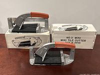 Two Mini Miki tile cutters placed side-by-side on a wooden surface, each with their original white boxes labeled 'No.3 Mini Miki Tile Cutter Made in Japan'.