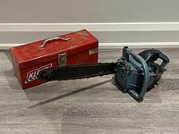 Photo of vintage blue Danarm chainsaw with black chain and handle, placed next to a red metal KLEIN toolbox on wooden floor.