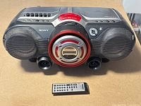 Front view of Sony boombox with remote placed in front on beige surface. Shows dual speakers, central CD player with red ring, tape deck window, controls on top and Sony logo.
