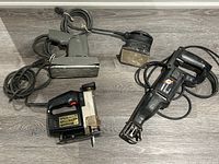 Top down view of two electric sanders (gray and black), scroll saw (black), and reciprocating saw (black) laid on wooden floor.