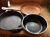 Three frying pans arranged side-by-side showing interiors with black non-stick, copper-toned metal, and black ceramic surfaces.