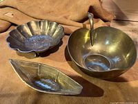 Lot of three old brass bowls visible together on cloth background. Visible are a leaf-shaped bowl, a round scalloped bowl with feet, and a round handled bowl.