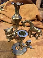Photo showing front view of four brass decorative items: giraffe figurine, vase, cat figurine, and candle holder.