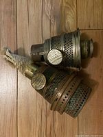 Two antique Aladdin lantern wick burner parts on wood floor. One has a rope wick attached. Both parts feature metal perforations and attached circular embossed Aladdin label.
