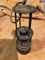 Antique brass wick lantern showing the main body, protective brass frame, and wick area, photographed against a brown fabric background.