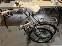 Folded mini folding bike in white color, showing handlebar, seat, small wheels, and folded frame.