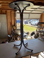 Side view of round black Ambrite bar table with curved metal legs, showing the table top with paint stains and the metal leg structure underneath.