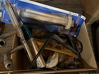 Box containing various tools and hardware including a packaged white table leg, metal hand tool, and wood items