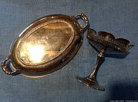 Oval silver-plated bowl with two handles and beaded rim next to a silver-plated pedestal candy dish with pierced scalloped edge, both showing original finish with age-appropriate wear and patina.