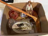 Photo showing box contents including vintage wooden hockey stick, glass jug, ceramic plaque, and figurines inside a cardboard box.