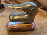 Shot of desk stapler featuring a brass duck head mounted on a light wood stapler body and dark wood base, placed on leather surface.