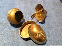 Three small brass items arranged on blue fabric: round vase, bowl with handle, mask-shaped container with removable lid.