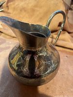 Brass water pitcher shown from the front and side angles, highlighting the curved handle and wide spout.